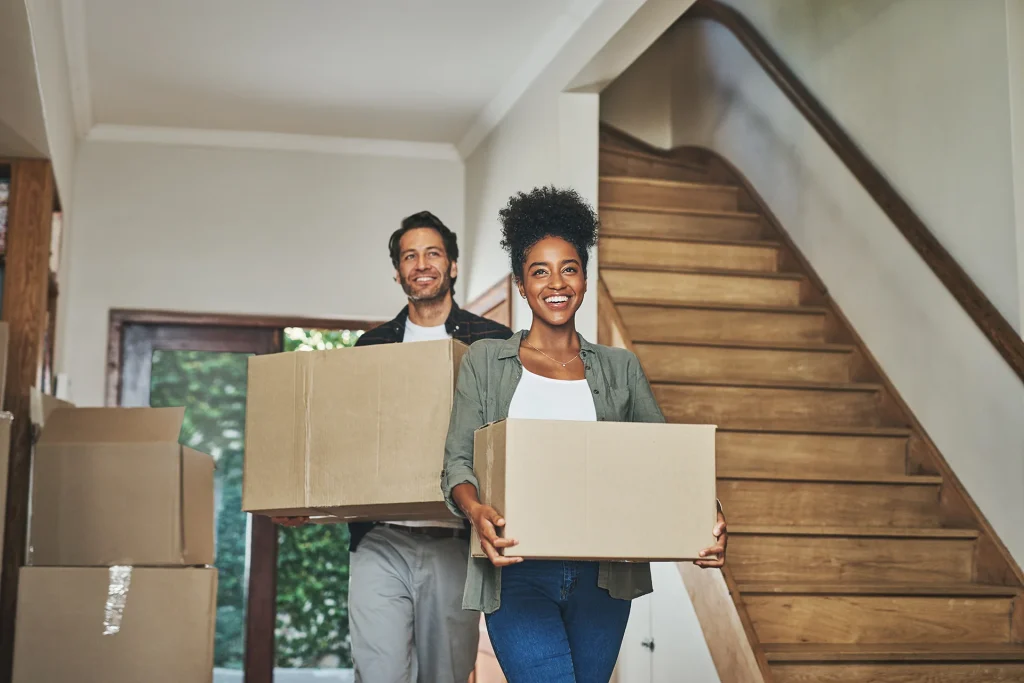 Smiling Couple Carrying Moving Boxes Into New Home